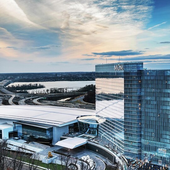 Aerial view of a modern, glass-fronted MGM hotel and casino by a river at sunset, with highways curving around it and a serene sky above, exuding luxury.