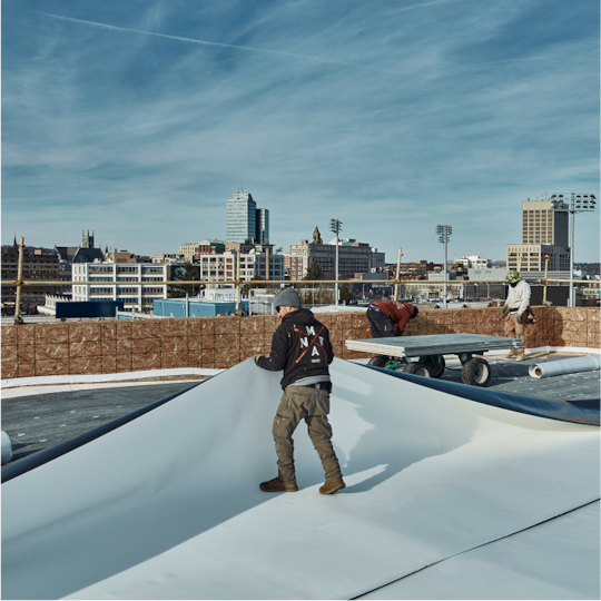 GAF Commercial Certified contractors installing a white TPO roofing membrane on a commercial rooftop with a city skyline in the background.