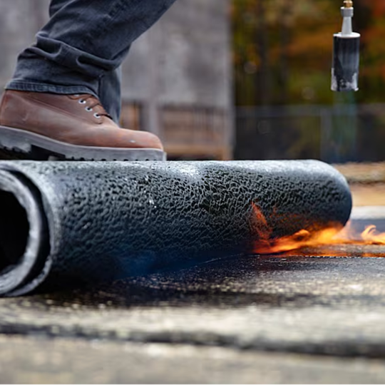 A GAF certified contractor torch-applying a roll of SBS modified bitumen membrane to a commercial roof deck.