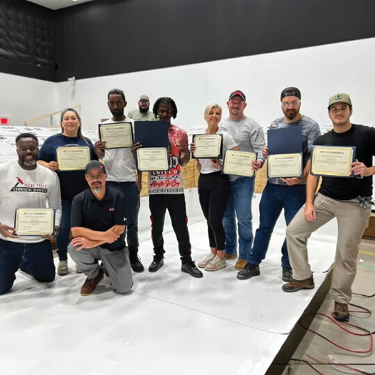 Group of GAF Roofing Academy graduates holding framed certificates in a training room.