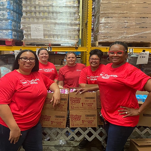 Six GAF employees wearing red shirts with a "Community Matters" logo stand smiling together in a warehouse where they are volunteering their time. 