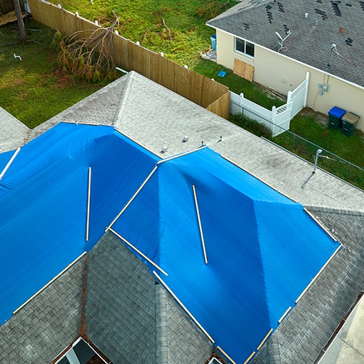 Aerial view of a home with a large blue tarp covering adamaged roof, illustrating the immediate need for restoration roofing services following a storm.