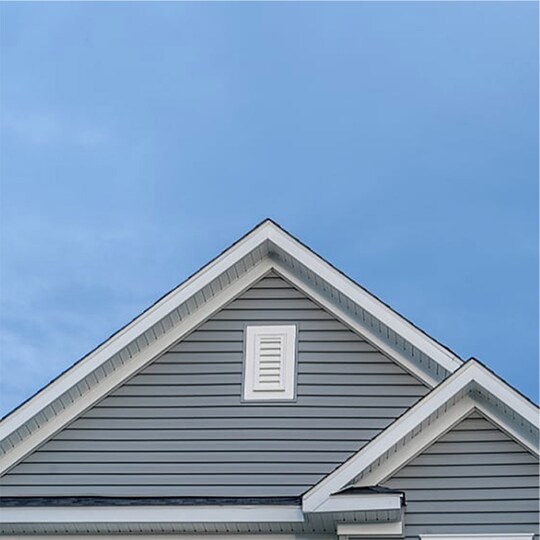  A close-up shot shows the gray siding and dark shingled roof of a house, highlighting a rectangular attic vent with a white frame centered in the main gable against a blue sky.