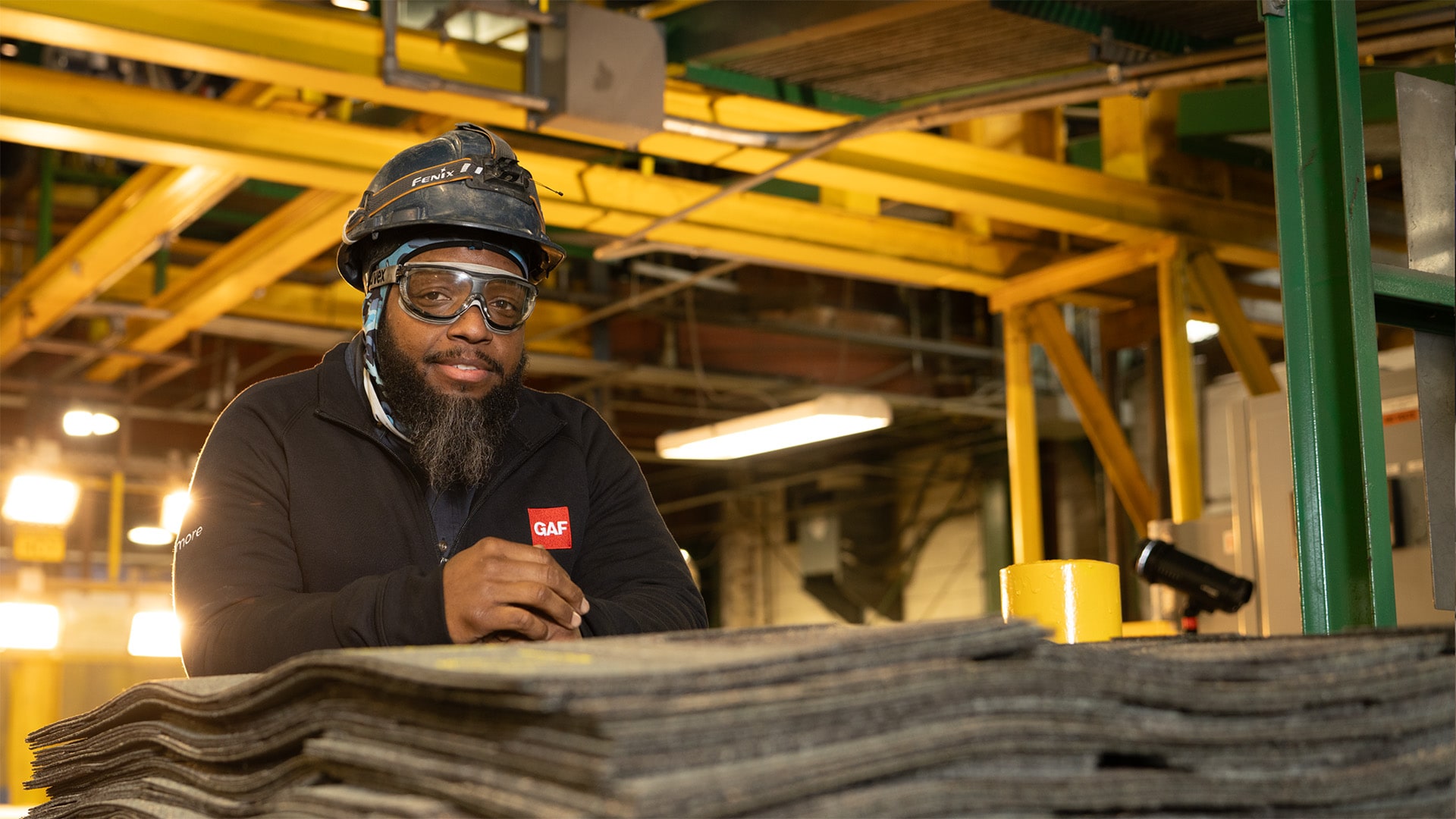 GAF employee in safety gear leaning over a stack of roofing shingles inside a manufacturing facility.