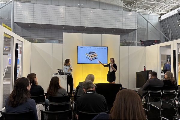 Two women presenting a technical diagram of a roofing assembly on a digital screen to an audience seated in a convention booth.