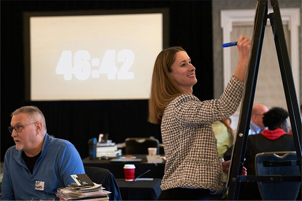 A photo of a woman writing on a large easel during a workshop, with a digital timer in the background displaying "46:42" and other attendees seated at tables.