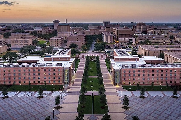 An aerial photo showcases a brick building complex with green landscaping under a sunset sky, likely demonstrating GAF’s roofing materials. The scene includes a wide view of other urban buildings in the background.