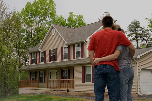 A homeowner couple is shown from behind, standing in a yard and embracing while looking at a two-story beige and maroon-trimmed house after a successful storm restoration project.