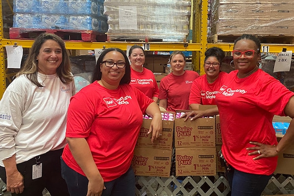 Six GAF employees wearing red shirts with a "Community Matters" logo stand smiling together in a warehouse where they are volunteering their time. 
