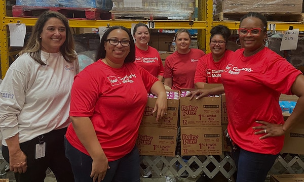 Six GAF employees wearing red shirts with a "Community Matters" logo stand smiling together in a warehouse where they are volunteering their time. 