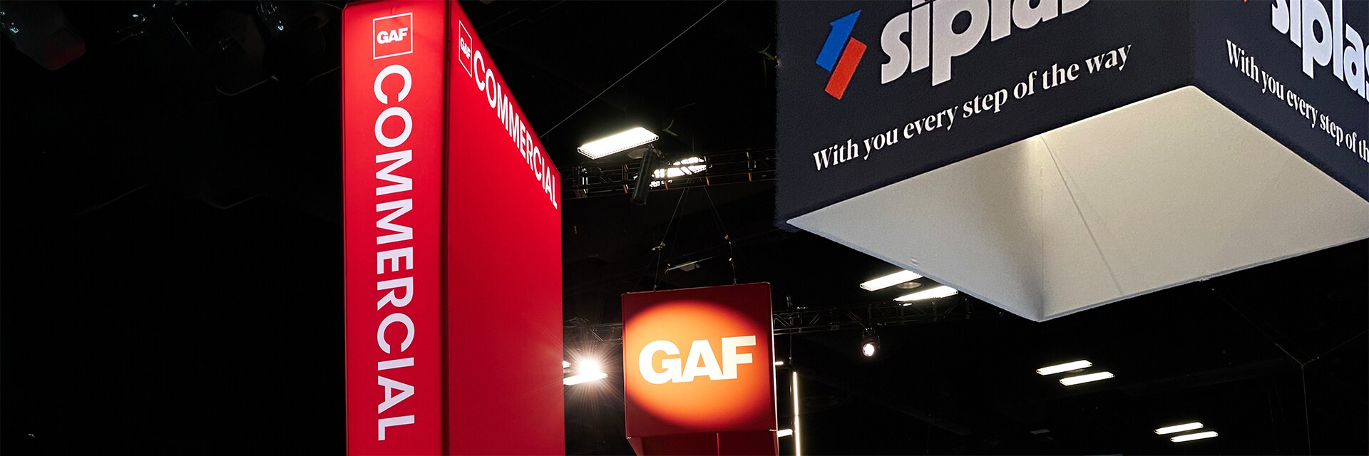 GAF Commercial and Siplast hanging signs at a AIA conference. Bright overhead lights illuminate the scene.