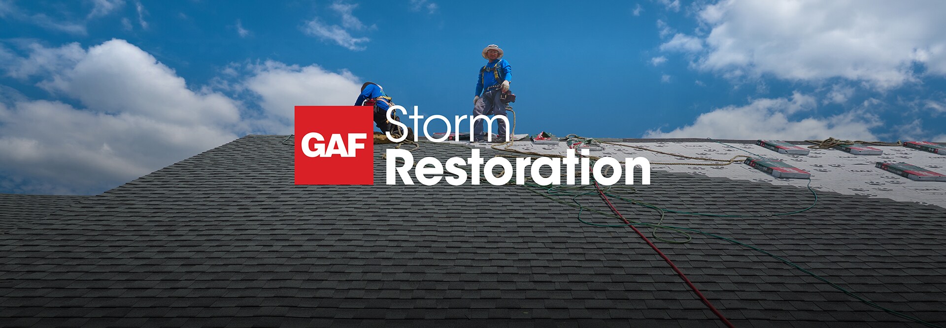 Two roofers working under a bright blue sky on a roof, with the GAF Storm Restoration logo overlaid on the foreground.