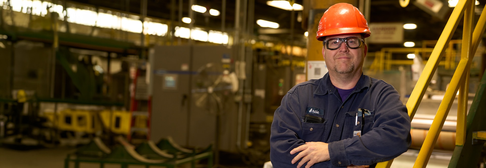GAF employee wearing an orange hard hat and safety glasses smiling with arms crossed inside a manufacturing plant.