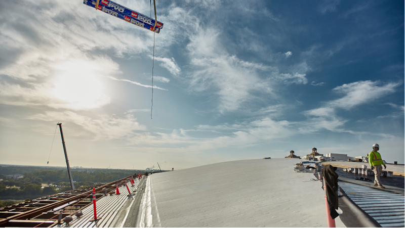 A crane lifts rolls of GAF EverGuard® PVC KEE membrane onto a large, white commercial roof under a bright, sunny sky.