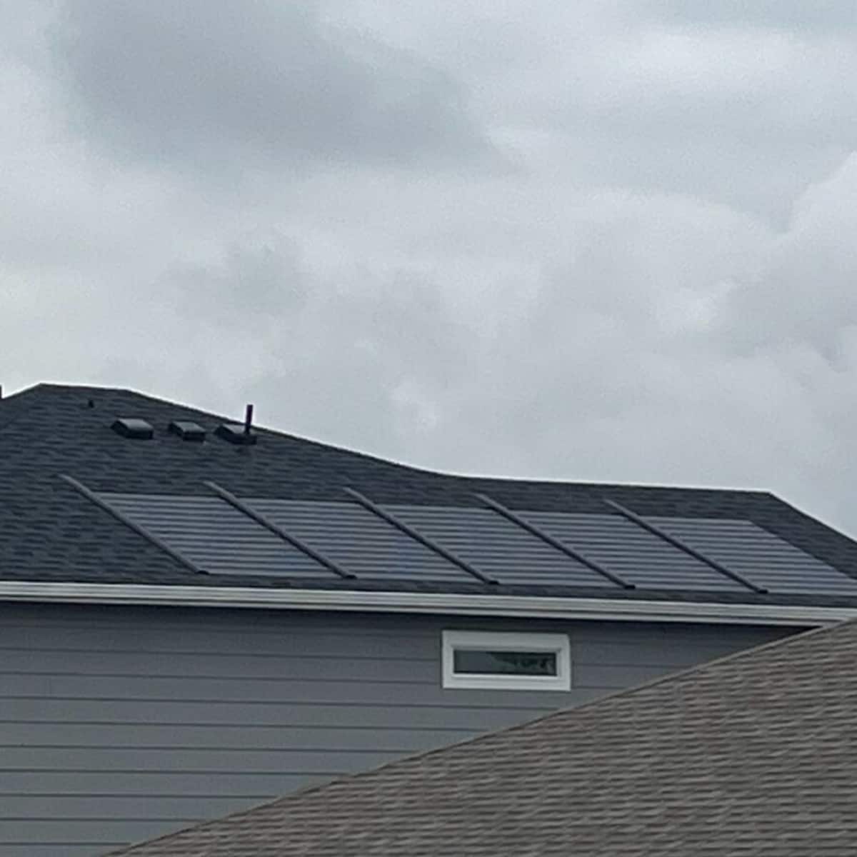 A close-up aerial view shows a residential roof with Timberline Solar® ES 2 shingles, with a suburban street and other houses visible in the background.