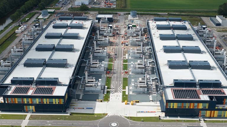 An aerial view of a large data center complex featuring two rectangular buildings with flat roofs covered in mechanical equipment and solar panels. 