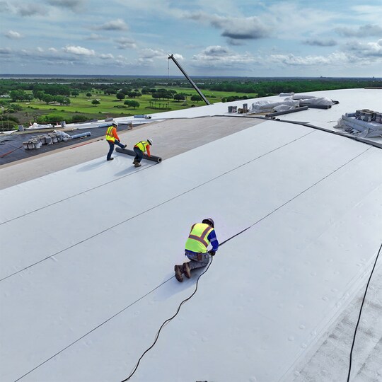 An aerial view of roofing contractors installing white PVC membrane on a large flat roof.