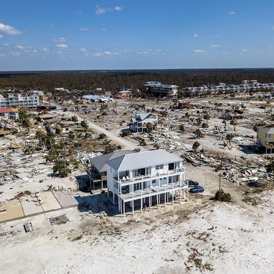 An elevated white house stands intact among widespread destruction from a storm. 