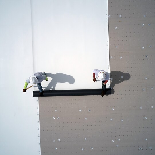 A top-down view of two roofing professionals unrolling a white membrane over a textured insulation layer on a flat roof.
