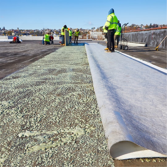 A worker unrolling a textured fleece-backed roofing sheet on a large commercial roof.
