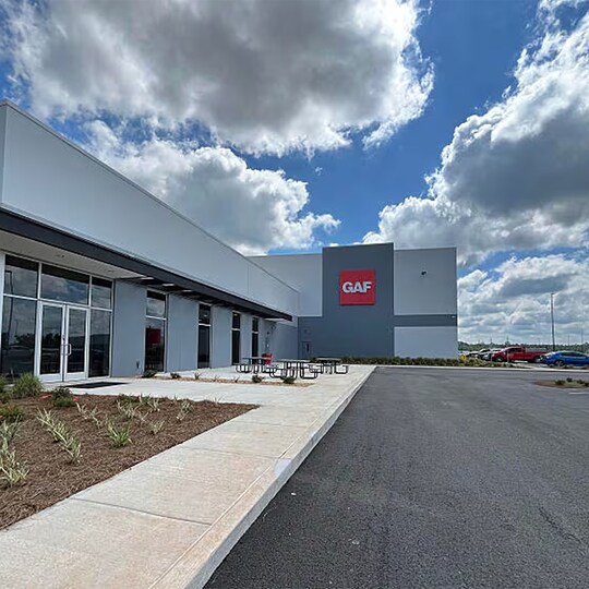 Exterior view of a modern GAF facility building with red branding, concrete walkways, and outdoor seating.