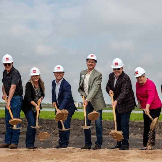 Six GAF and community leaders wearing white hard hats holding gold shovels at a facility groundbreaking ceremony.