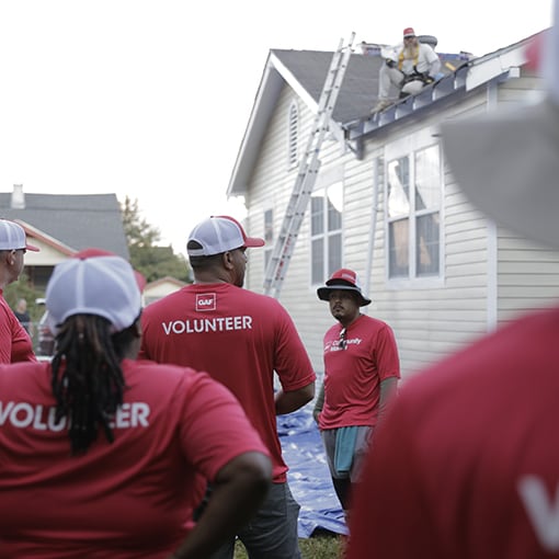 A group of GAF volunteers in red shirts with the word "VOLUNTEER" on the back are gathered on the ground while one worker in a safety harness is visible on the roof of a house.