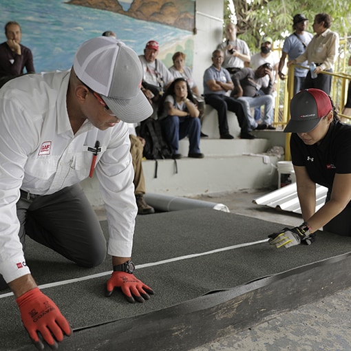 Two volunteer roofers are crouched down and working together to measure and prepare a piece of roofing material.