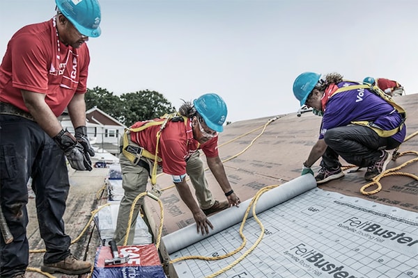Three volunteers in safety gear and hard hats are working on a residential rooftop, installing a layer of GAF FeltBuster synthetic roofing felt.