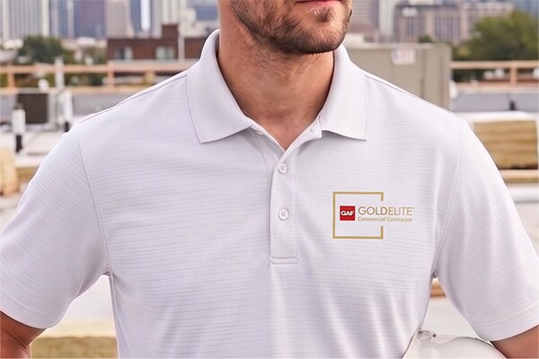 A GAF GoldElite Commercial Certified contractor in a white polo shirt standing on a rooftop holding a hard hat.