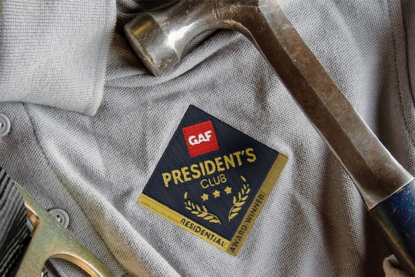 GAF President's Club patch on a contractor's shirt with construction tools nearby.