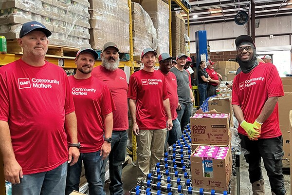 GAF employees wearing red shirts with a "Community Matters" logo stand smiling together in a warehouse where they are volunteering their time. 