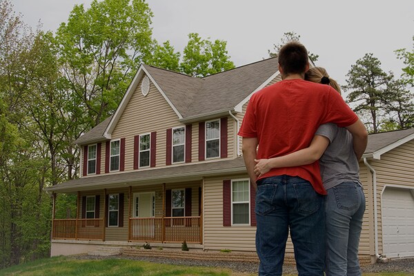A homeowner couple is shown from behind, standing in a yard and embracing while looking at a two-story beige and maroon-trimmed house after a successful storm restoration project.