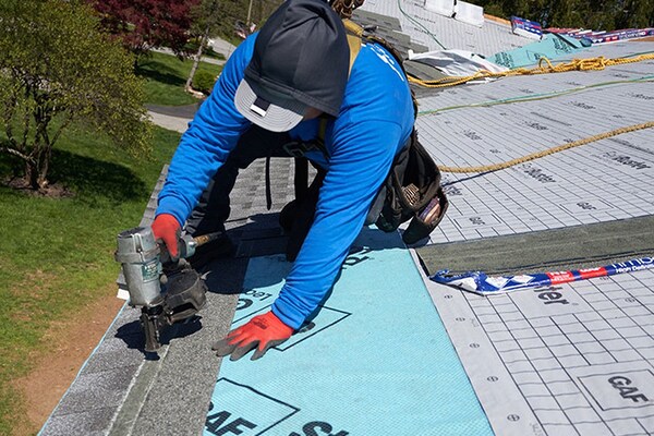 Two contractors working to install new shingles on a roof over a blue underlayment.