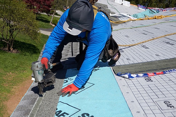 Two contractors working to install new shingles on a roof over a blue underlayment.