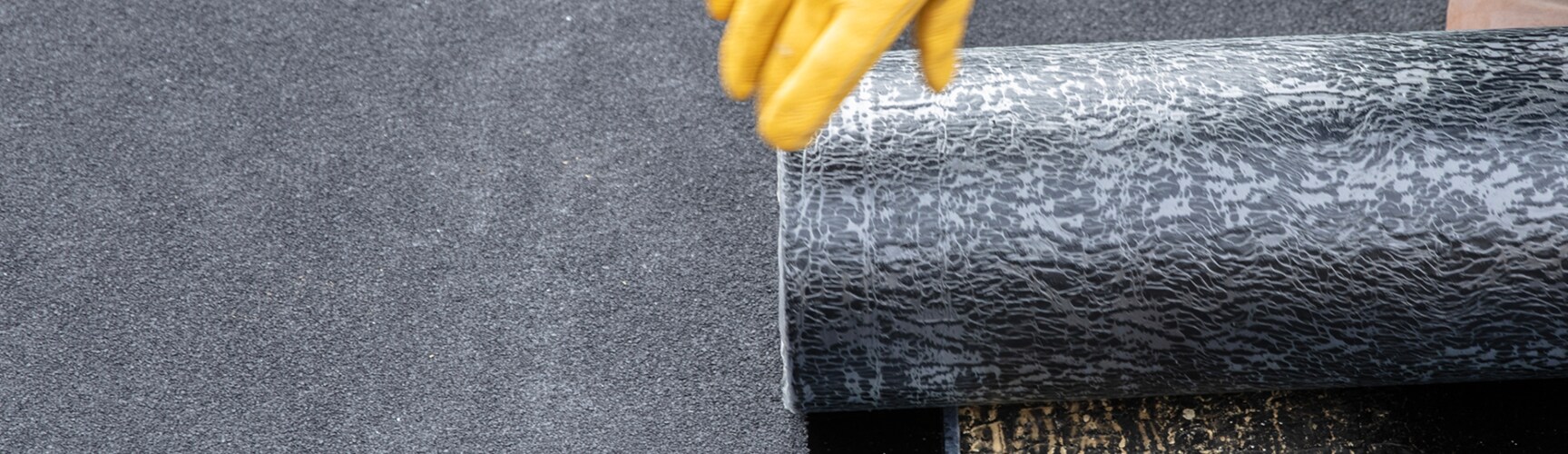 Gloved hands of a GAF certified contractor unrolling a dark, granular-surfaced modified bitumen roofing membrane.