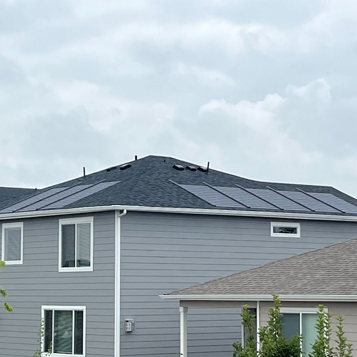 A two-story residential house with gray siding and a charcoal shingled roof has solar tiles from GAF Energy mounted. 