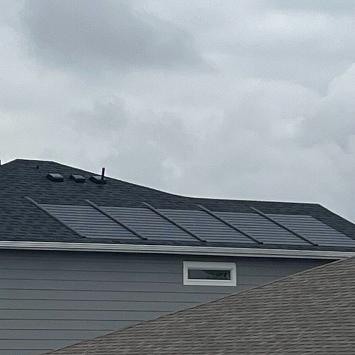 A close-up aerial view shows a residential roof with Timberline Solar® ES 2 shingles, with a suburban street and other houses visible in the background.